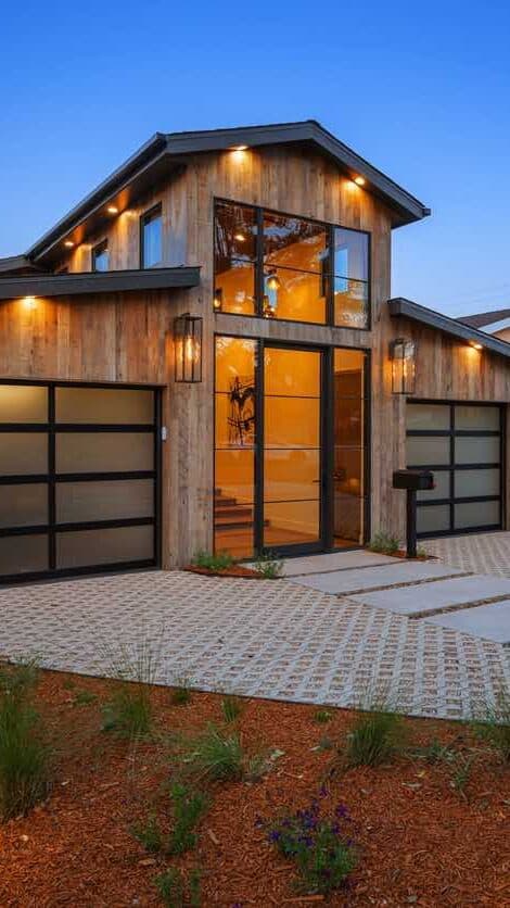 Modern barndominium-style home with wood siding, large glass entry, and two garage doors, with landscaped driveway and pathway.